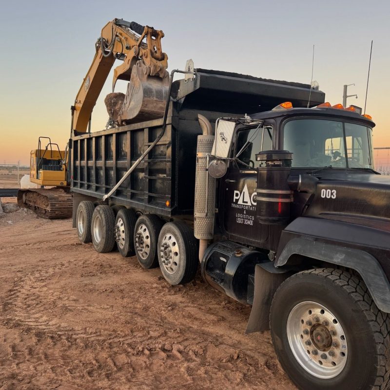 Excavator loads rocks into heavy-duty dump truck at construction site during sunset, earthmoving and hauling.