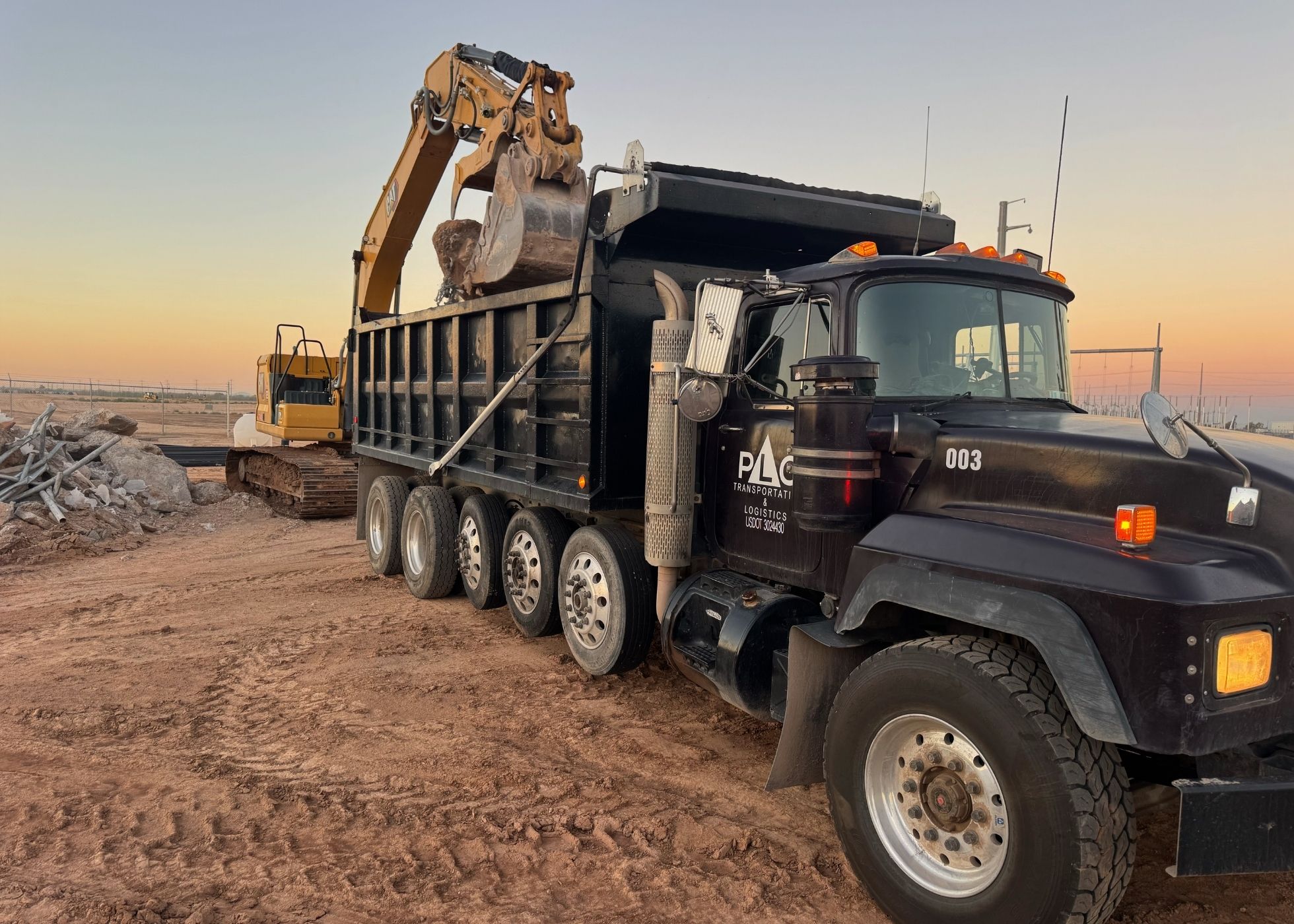 Excavator loads rocks into heavy-duty dump truck at construction site during sunset, earthmoving and hauling.