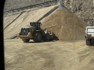 Front-end loader moving sand at quarry under conveyor stacker, large gravel pile and dump truck at mining site.