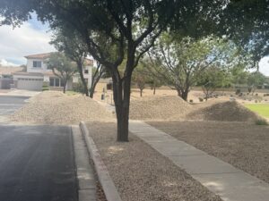 Large gravel piles for xeriscape landscaping along desert suburban street with trees and sidewalk near a two-story house