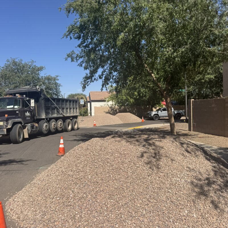 Dump truck delivering landscaping gravel on residential street, traffic cones and rock piles by sidewalk.