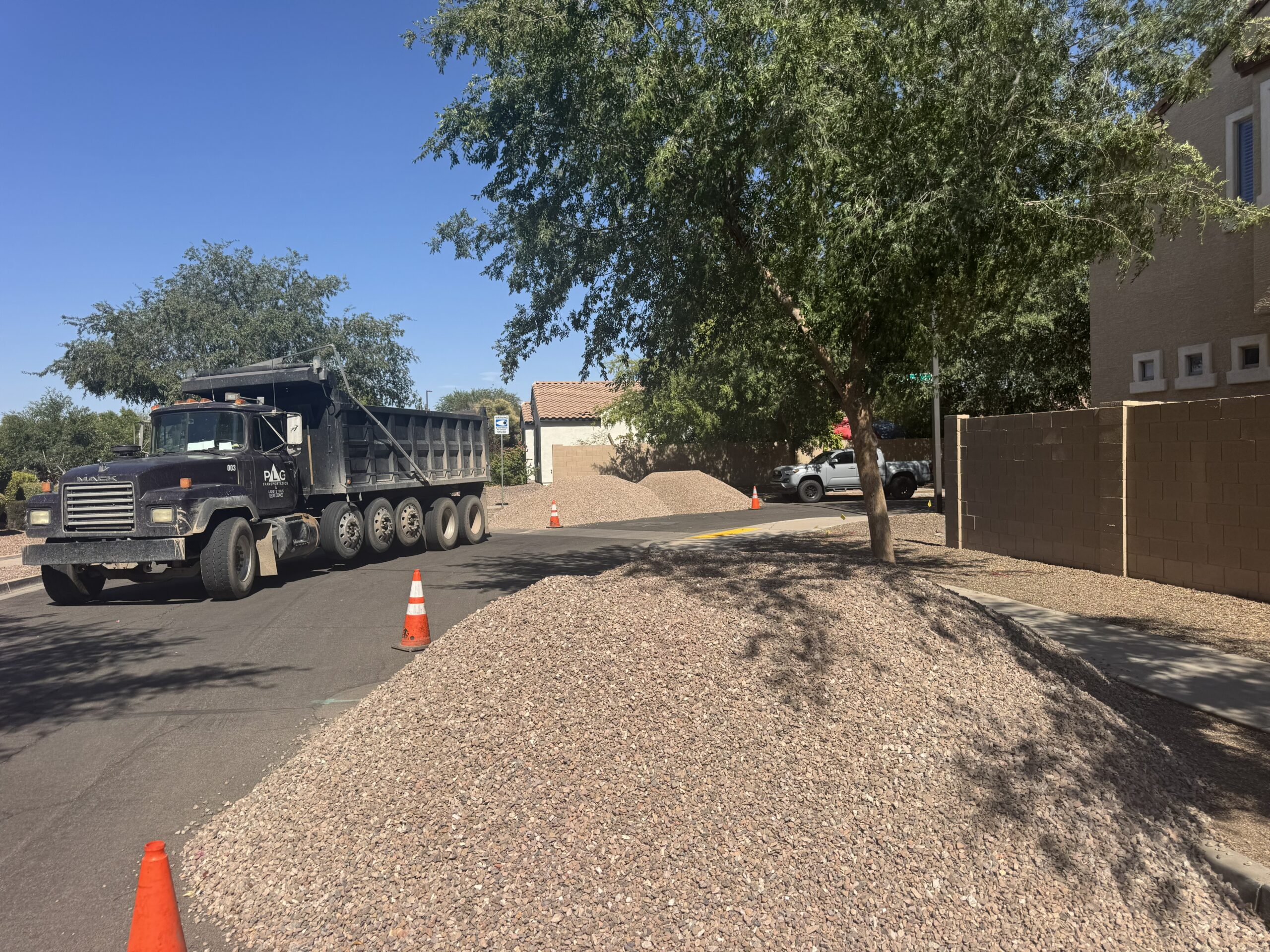 Dump truck delivering landscaping gravel on residential street, traffic cones and rock piles by sidewalk.