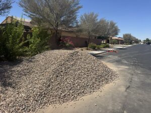 Curbside landscape rock delivery: large gravel pile on suburban desert street with trees and safety cones under blue sky.