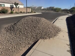 Landscaping gravel delivery pile on suburban cul-de-sac curb and sidewalk in a desert neighborhood under clear blue sky.