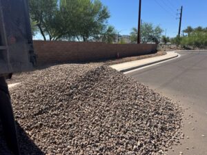 Dump truck delivers landscape gravel on suburban street near curb and sidewalk in desert neighborhood.