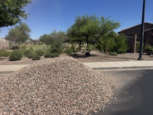 Large pile of landscaping gravel delivered by curb near desert xeriscape park and trees on a sunny suburban street.