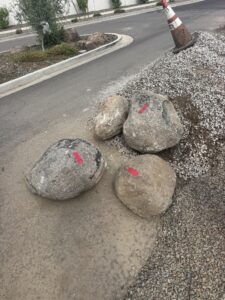 Boulders with red tape beside gravel pile and traffic cone on suburban street landscaping construction site.