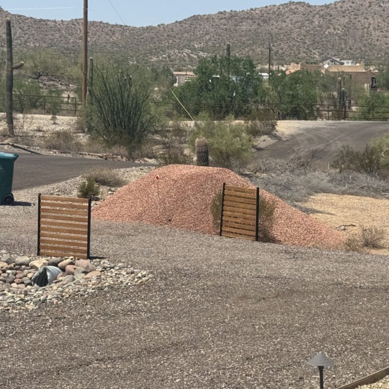 Desert landscaping scene with red gravel pile, saguaro cacti, and mountain backdrop on an Arizona residential road.