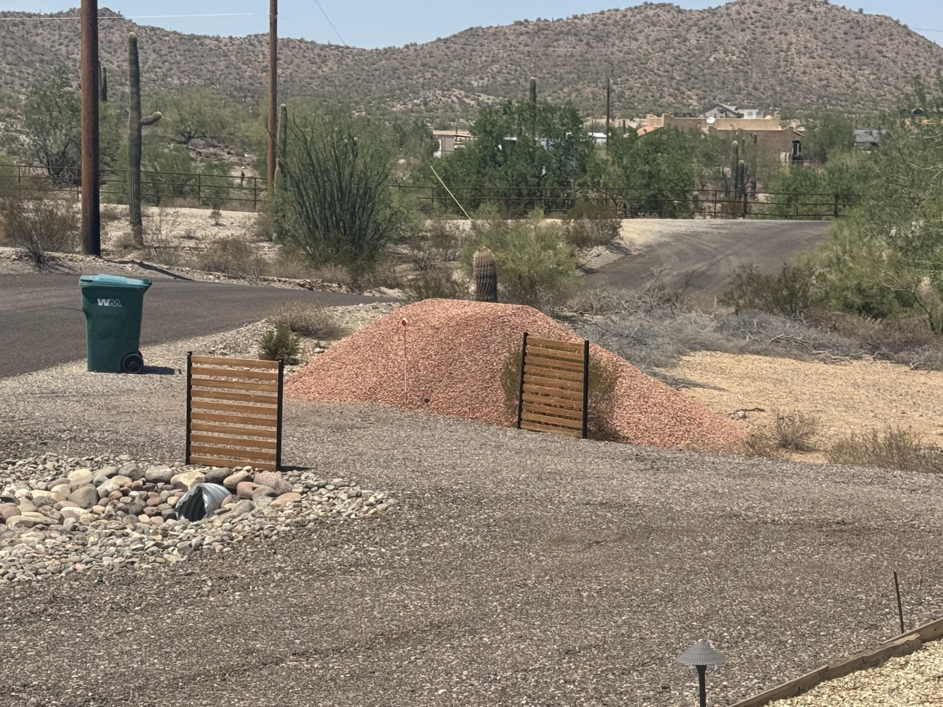 Desert landscaping scene with red gravel pile, saguaro cacti, and mountain backdrop on an Arizona residential road.