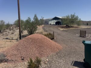 Arizona desert home with carport and SUV, large pile of red landscape rock on gravel driveway, xeriscape yard.