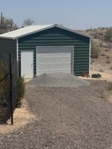 Green metal garage with roll-up door and side entry, gravel pile in front, Arizona desert landscape.