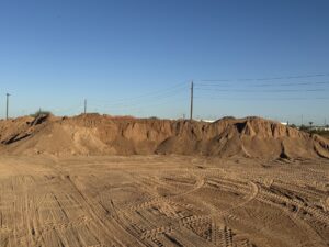 Sand and soil stockpiles at construction site with tire tracks, blue sky, and power lines in a desert landscape.