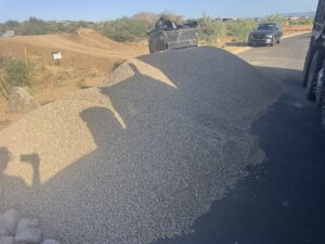 Gravel delivery on asphalt driveway at desert construction site, large rock pile beside dumpster and parked car.