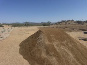 Desert landscaping site with large gravel mound, xeriscape preparation and distant mountains under clear blue sky.