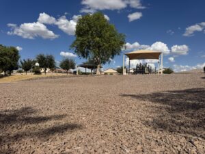 Covered playground with shade canopy in desert park, gravel landscaping, trees, and blue sky with clouds