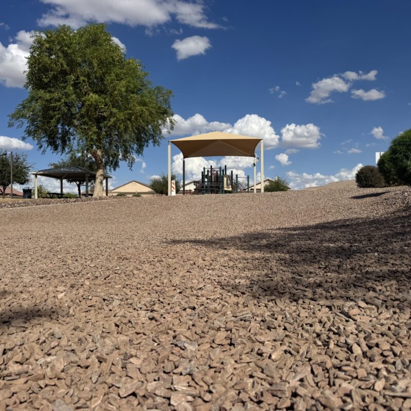 Desert park playground with shade canopy and gazebo, gravel landscape, green trees, and blue sky with clouds