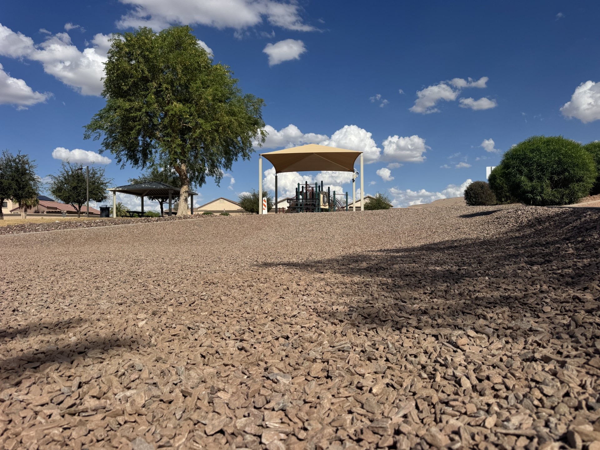 Desert park playground with shade canopy and gazebo, gravel landscape, green trees, and blue sky with clouds