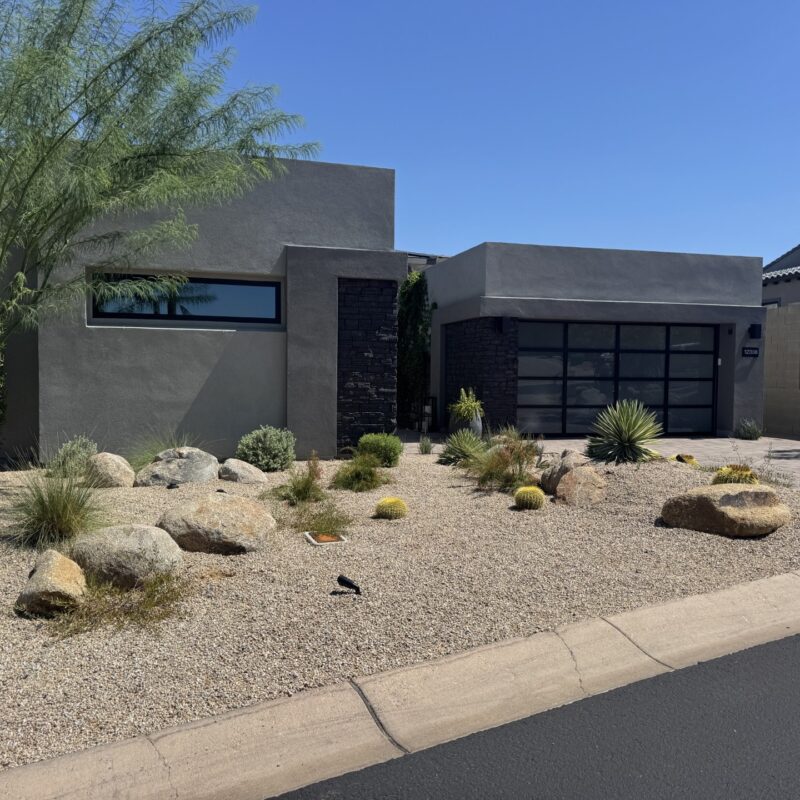 Modern desert home with xeriscape front yard, cacti and rocks, gray stucco, glass garage door under clear blue sky.